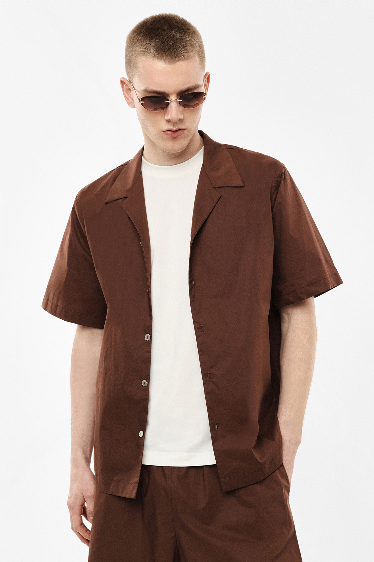Young man wearing brown short-sleeved shirt and matching shorts over a white T-shirt, wearing sunglasses