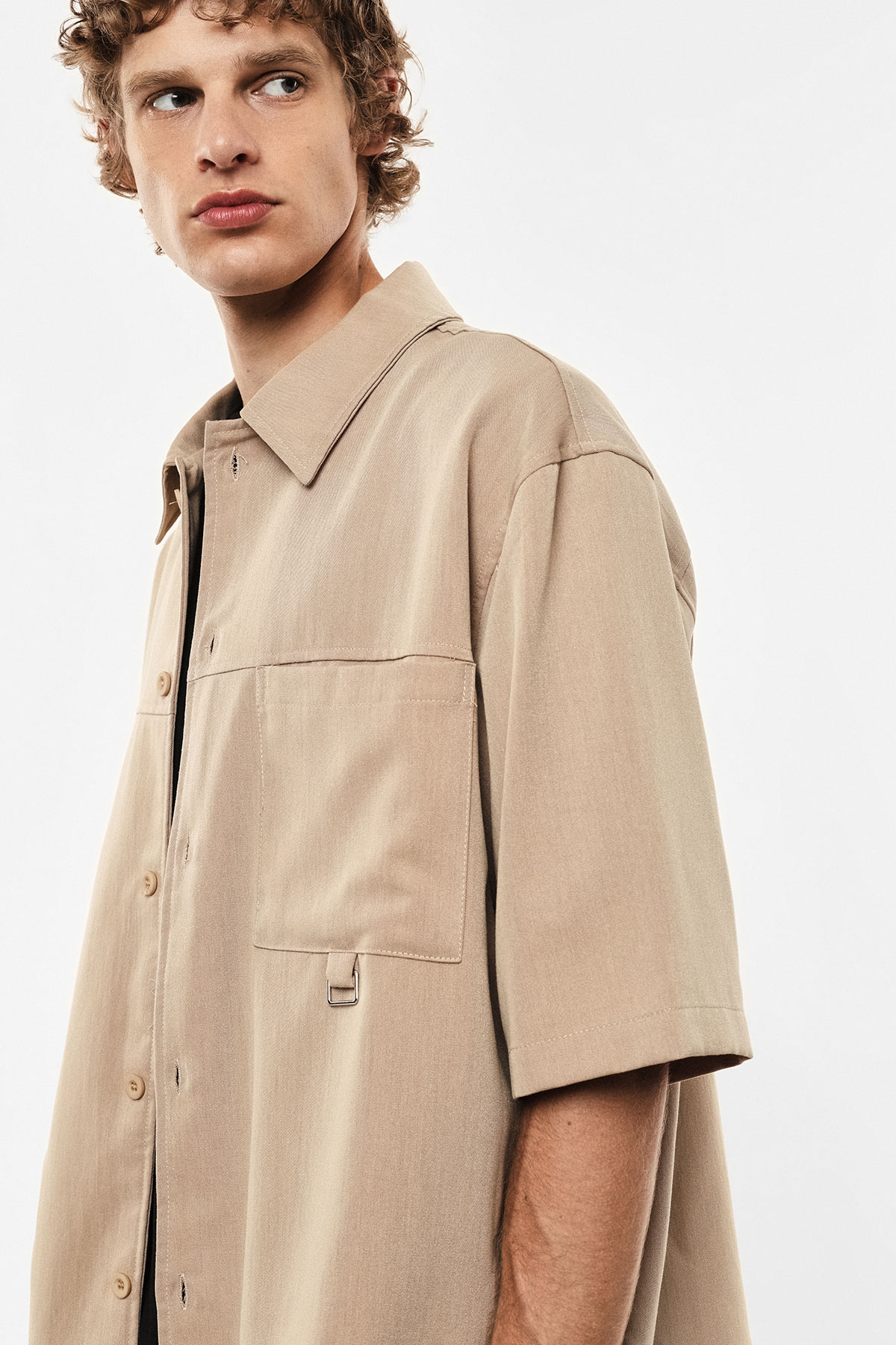 Young male model wearing a beige short-sleeve shirt with chest pockets, looking left against a white background