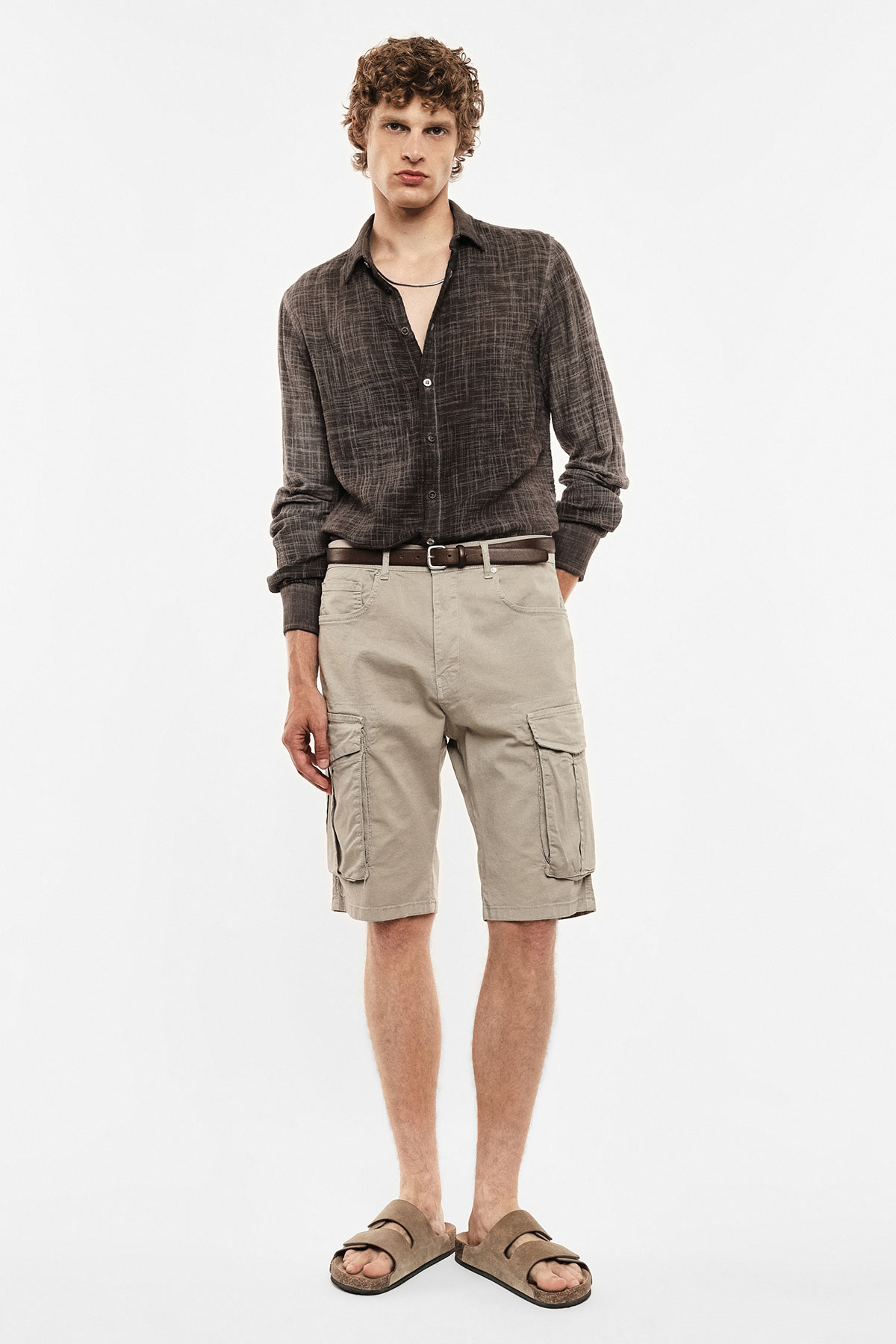 Male model wearing dark brown textured shirt, beige cargo shorts with side pockets, brown belt and suede sandals on white background