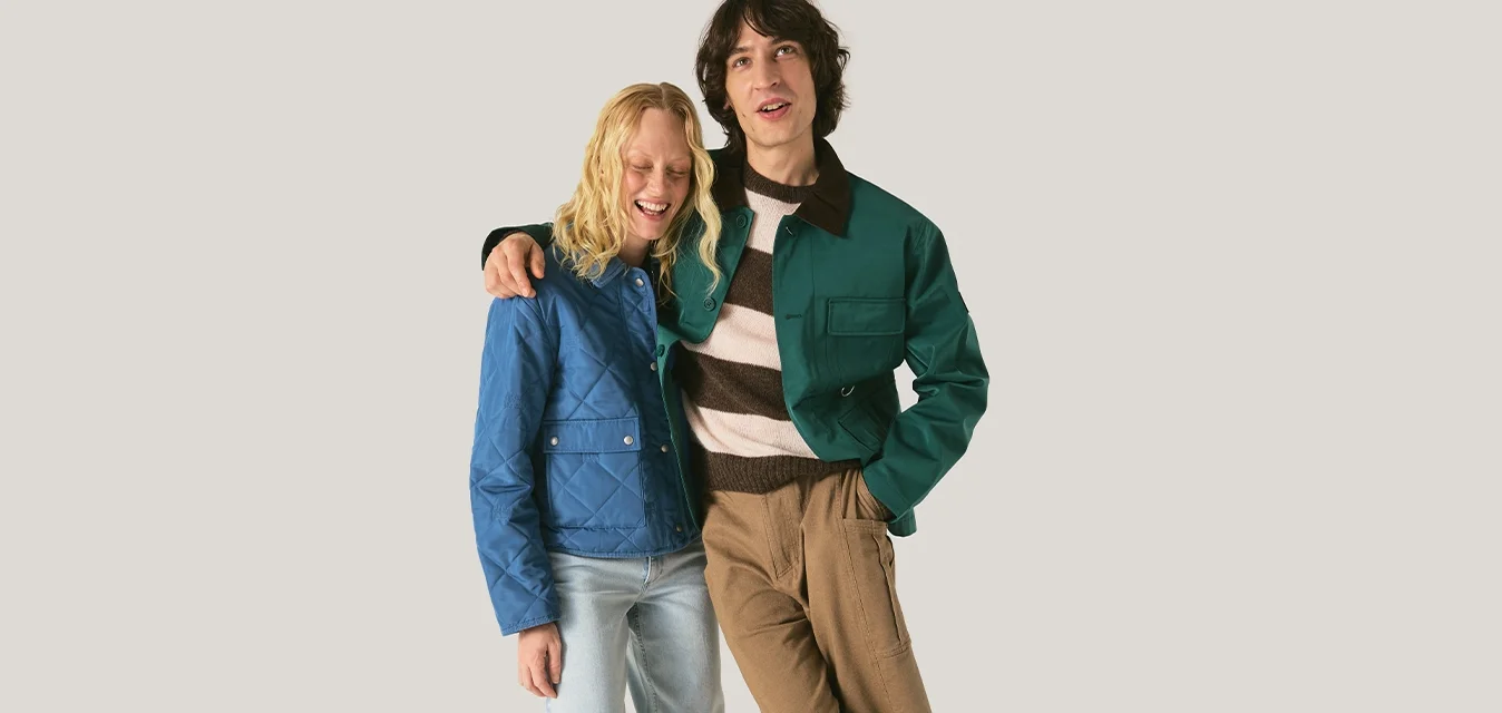 Due giovani adulti stanno vicini; uno indossa una giacca blu e jeans chiari, l'altro una giacca verde e una camicia a righe con pantaloni marroni.