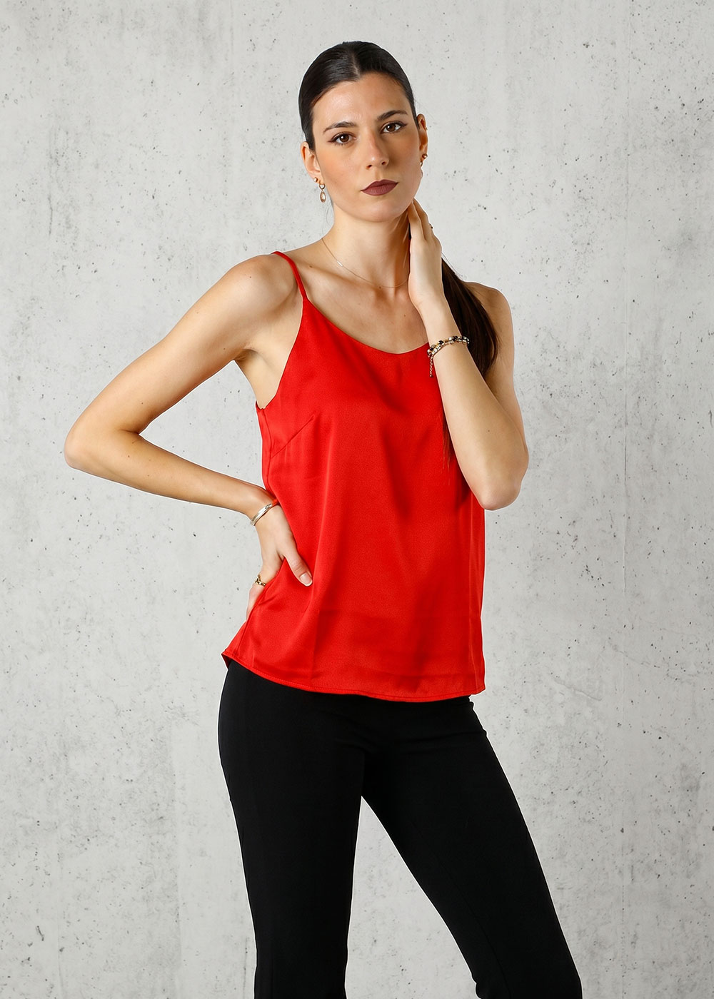 Female model wearing a red camisole top and black trousers, standing against a light concrete wall with one hand on her hip
