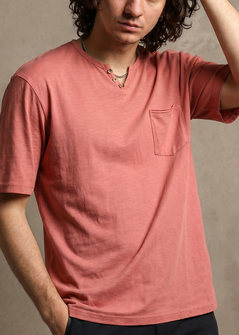 Man wearing faded rose short-sleeve pocket T-shirt with henley button neckline and chain necklace, hand in pocket, cropped torso view