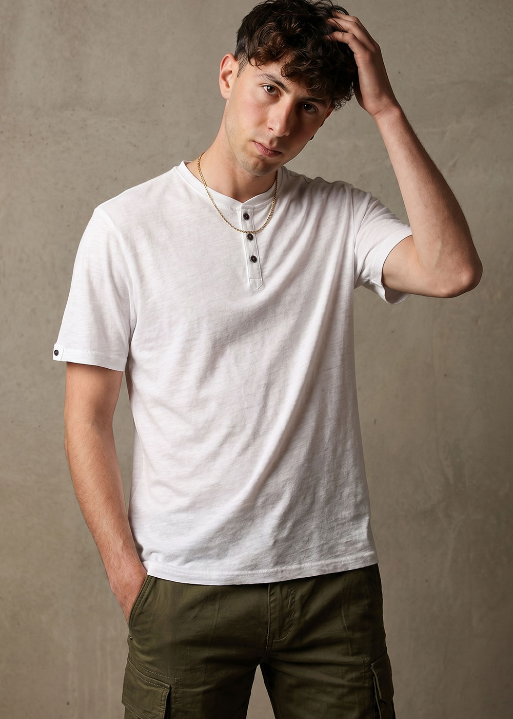 Young man in white henley t-shirt and olive cargo trousers, hand in pocket and touching hair against a concrete wall