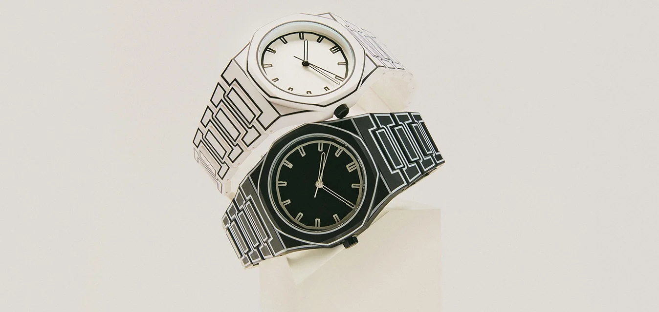 Un paio di orologi da polso, uno nero e uno bianco, con cinturini a motivo geometrico, esposti su un supporto bianco su uno sfondo chiaro.