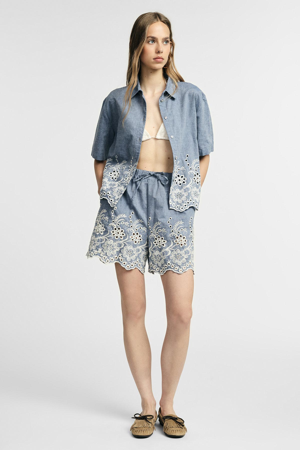 Woman wearing blue chambray embroidered short-sleeve shirt and matching scalloped shorts over white bikini top, tan moccasin shoes