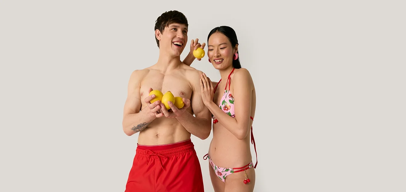 Giovane uomo con boxer da bagno rossi e donna in bikini floreale che tengono giocoso anatroccoli di gomma gialli su uno sfondo chiaro uniforme.