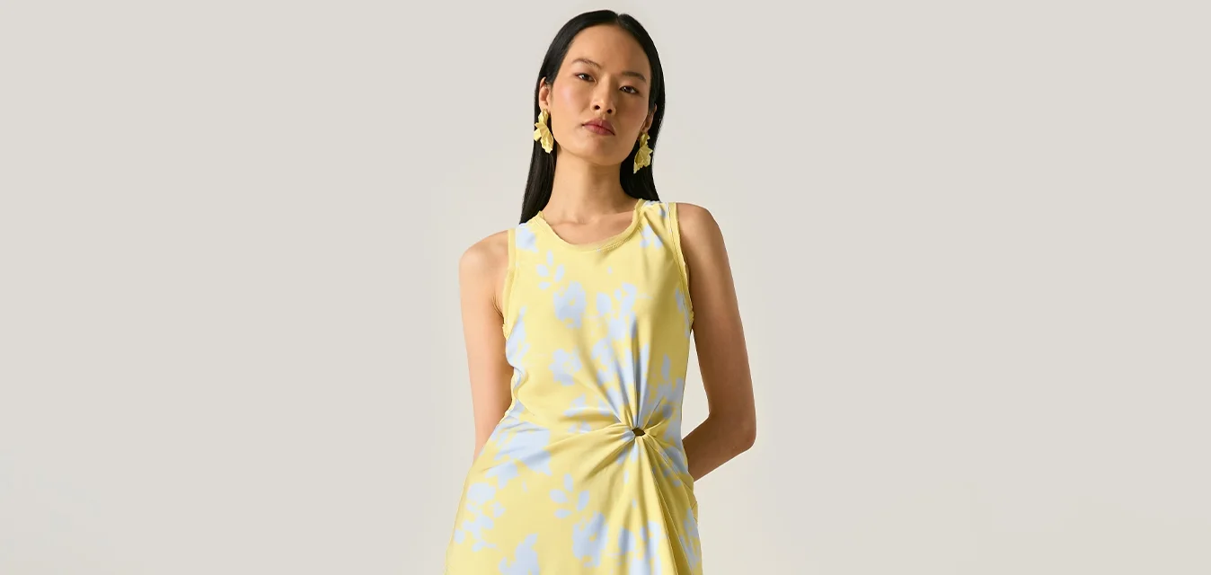 Donna con abito floreale giallo e blu senza maniche con dettaglio a torsione laterale, in piedi accanto a una pianta di monstera in vaso su sfondo semplice.