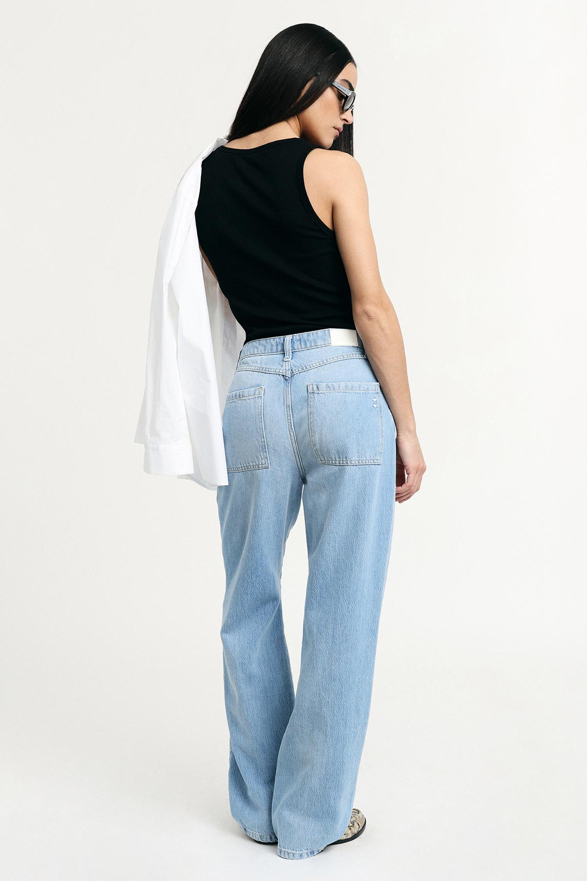 Woman model seen from back wearing a black tank top, light-blue wide-leg jeans and sunglasses, white shirt draped over shoulder
