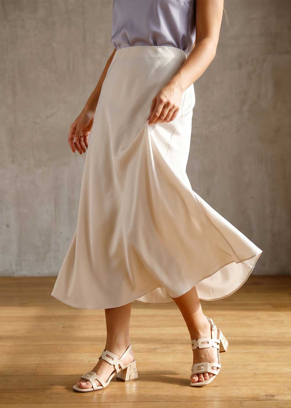 Lower half of woman modelling cream satin midi skirt and beige block-heel sandals on wooden floor