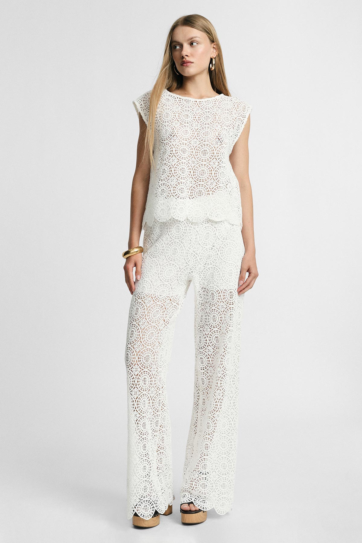 Full-length photo of a model wearing a white crochet lace two-piece top and wide-leg trousers, gold jewellery and platform sandals, neutral background