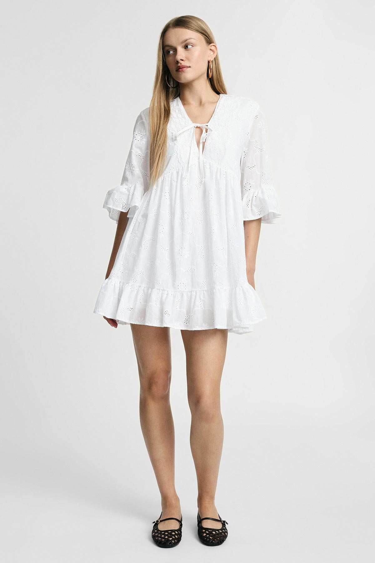 Woman wearing white eyelet babydoll dress with tie-front and ruffled hem, hoop earrings and black flats on plain background