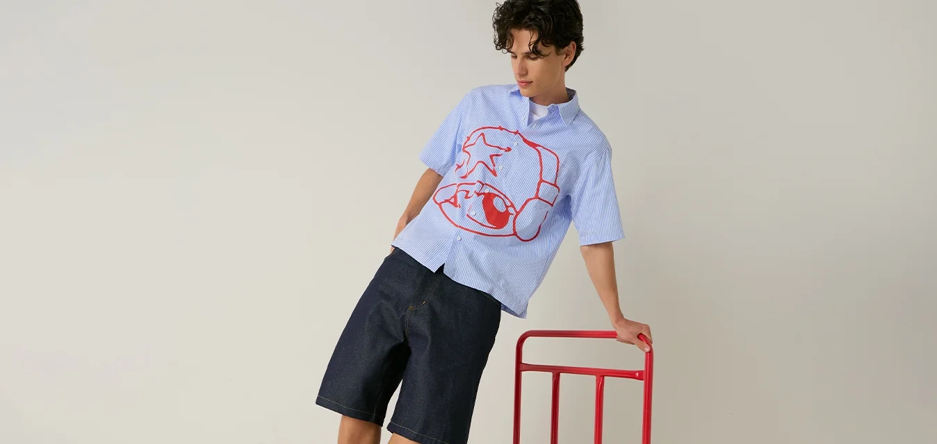 Giovane uomo che si equilibra su un carrello piattaforma rosso, indossa una maglietta blu con grafica rossa, pantaloncini neri, calzini bianchi e scarpe da ginnastica rosse, su sfondo semplice.