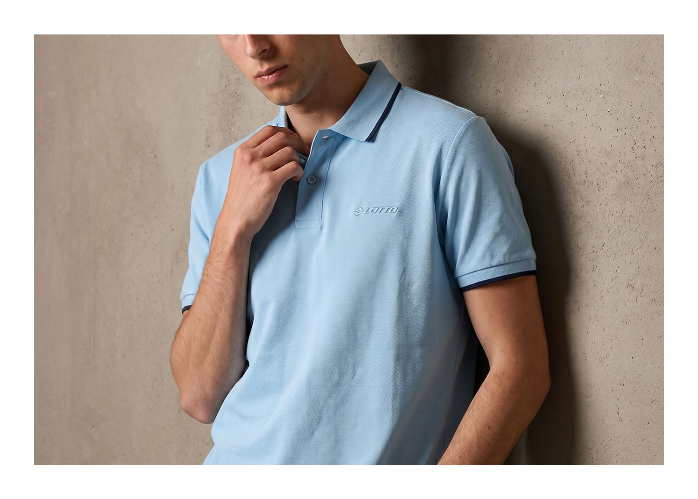 Cropped image of man in light blue polo shirt with Lotto logo, hand adjusting collar while leaning on a concrete wall