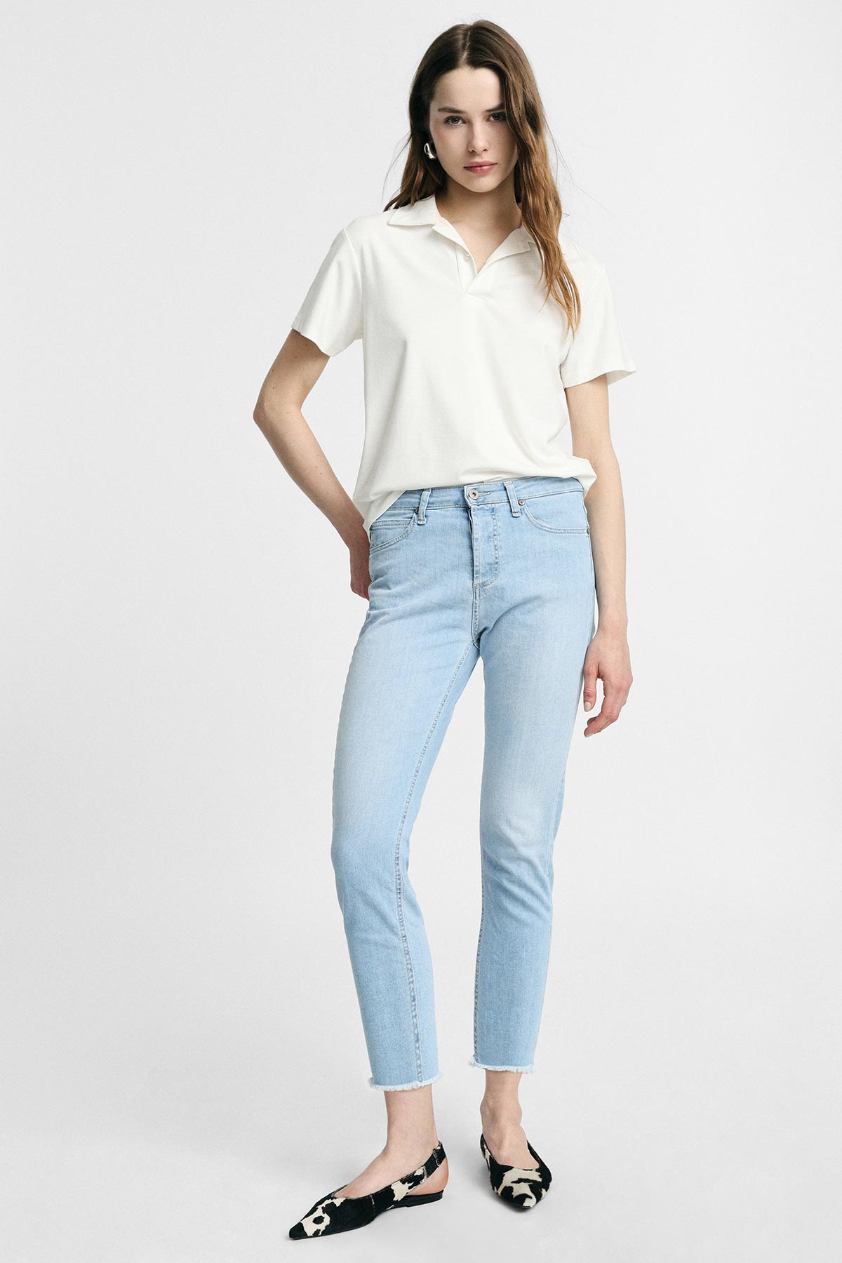 Woman wearing white polo shirt and light blue skinny jeans with black patterned slingback flats, standing hand on hip on plain background