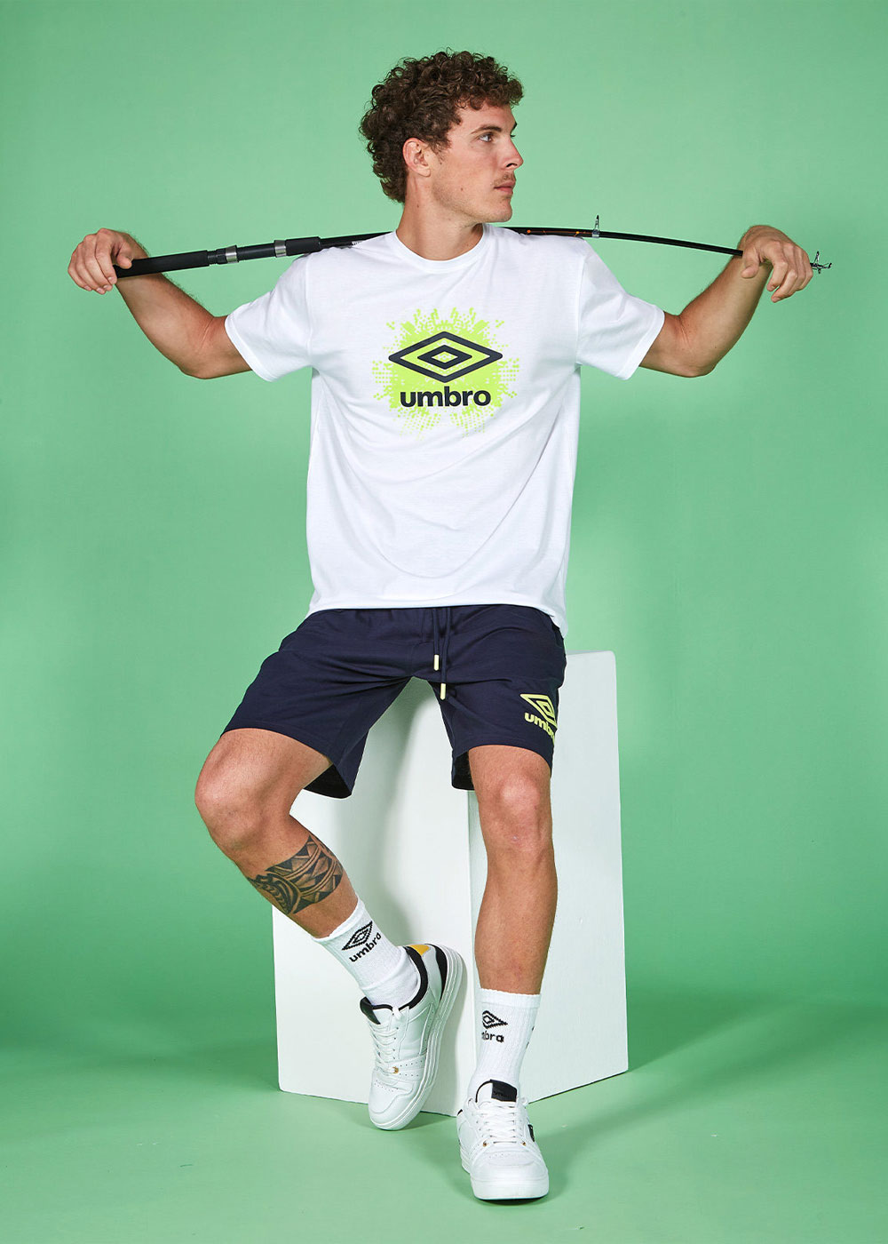 Man in white Umbro t-shirt and navy shorts sitting on a white block, holding a fishing rod across his shoulders against a green background