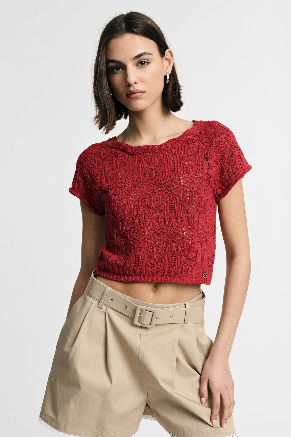 Female model wearing a red knitted short-sleeve cropped top and high-waist beige belted shorts against a white background