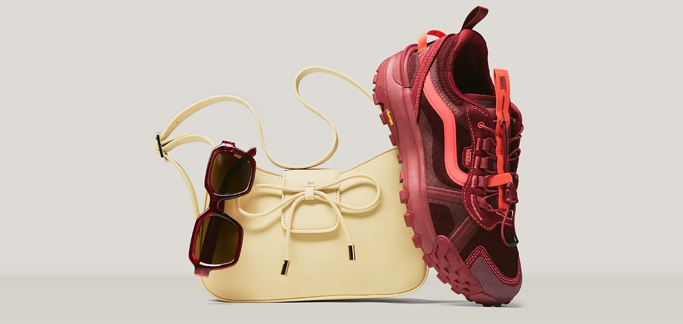 Borsa beige con dettaglio a fiocco, occhiali da sole scuri appesi alla tracolla e una scarpa sportiva rossa e rosa appoggiata alla borsa.