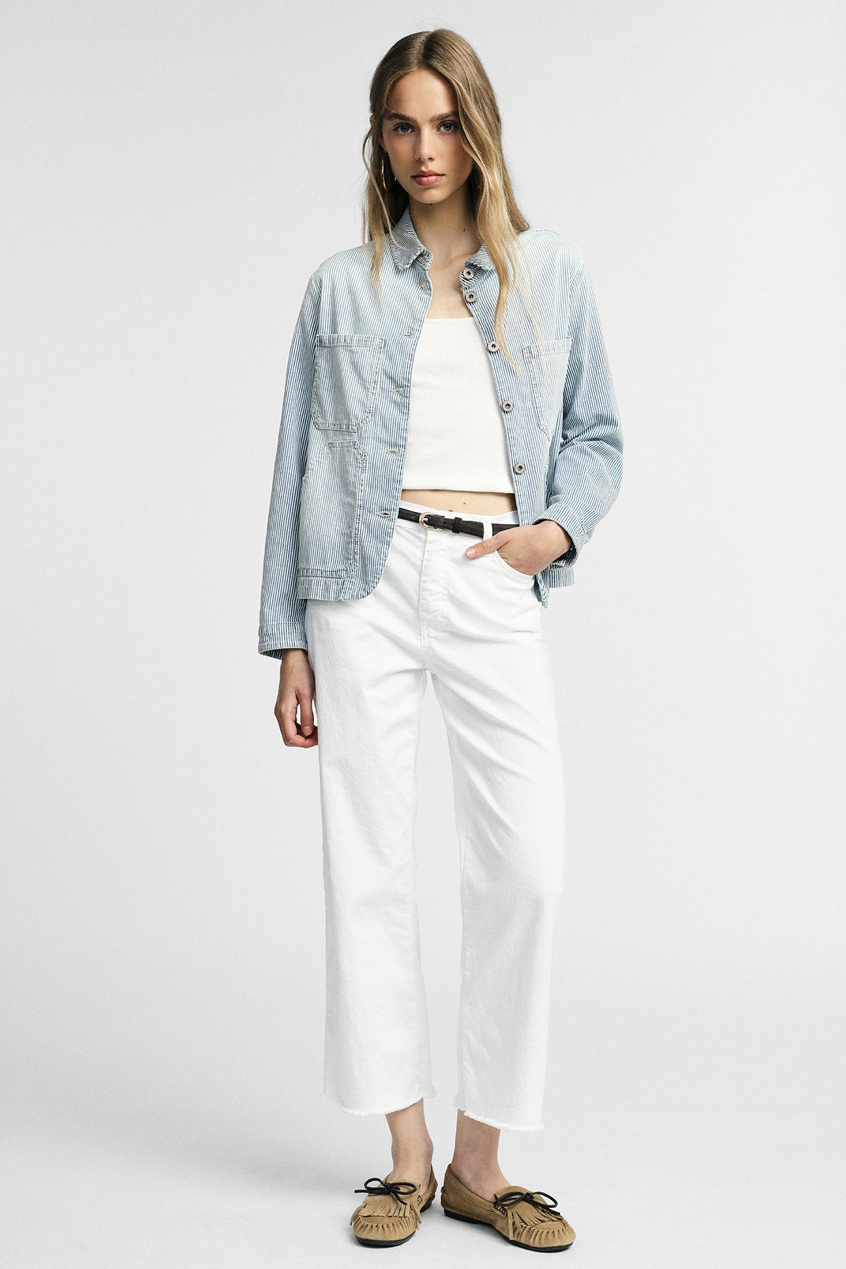Woman modelling casual outfit: striped light-blue jacket, white cropped top, high‑waist wide‑leg white jeans and brown moccasins, hand in pocket
