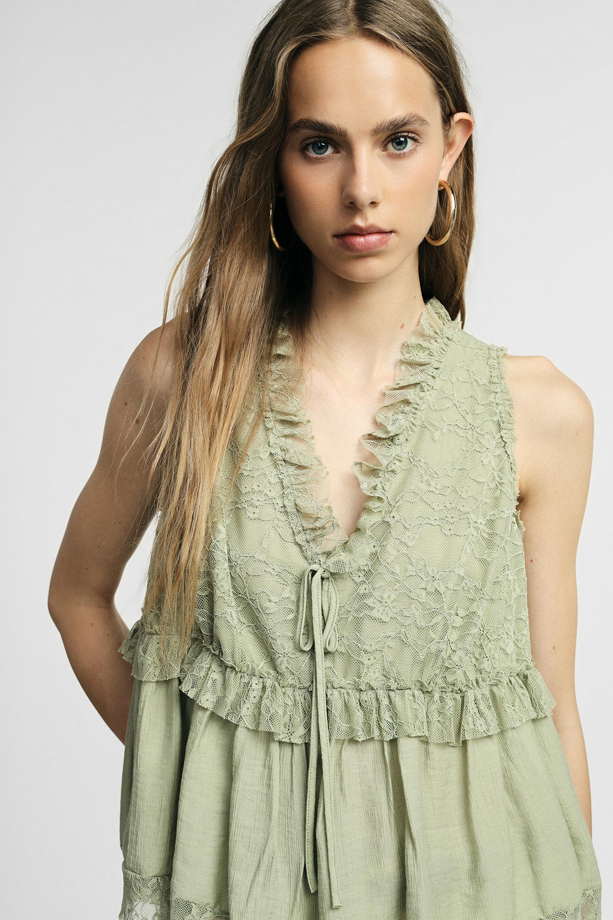Close-up of a young woman wearing a sage green sleeveless lace top with ruffled V-neck and hoop earrings against a neutral background