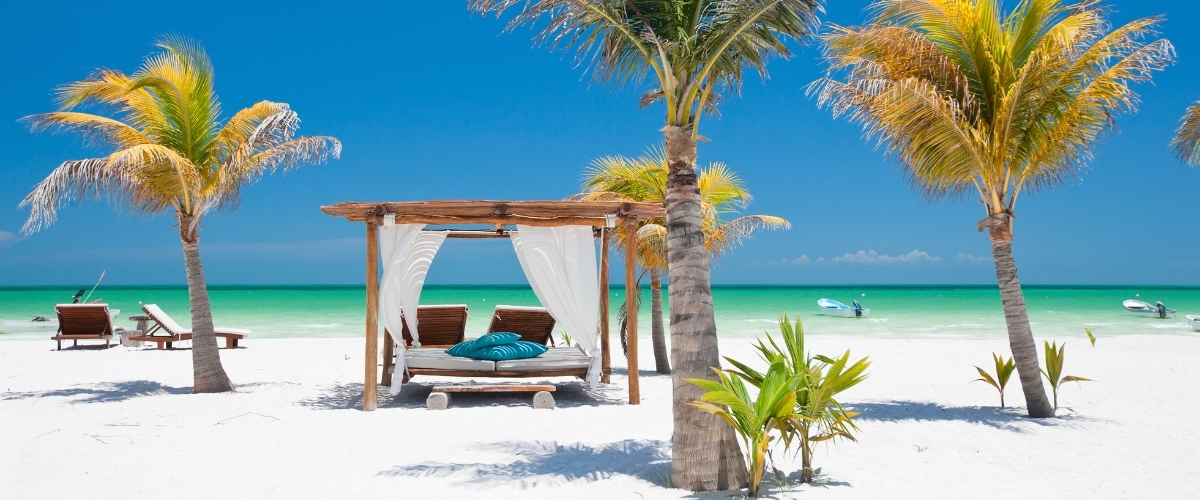 White-sand tropical beach with palm trees and a wooden cabana bed draped in white curtains and blue cushions facing the turquoise sea under a clear blue sky