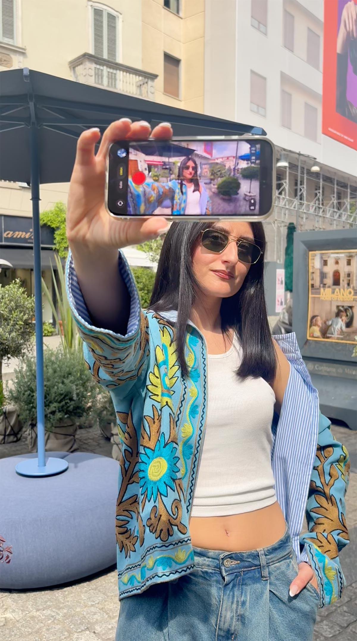 Young woman in sunglasses taking a smartphone selfie in an urban plaza, wearing a floral patterned jacket, white crop top and jeans
