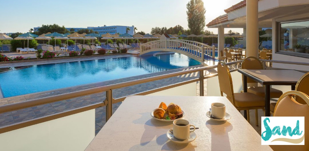 Table with coffee and pastries overlooking a sunlit resort pool, arched bridge and sun loungers with parasols — Sand logo