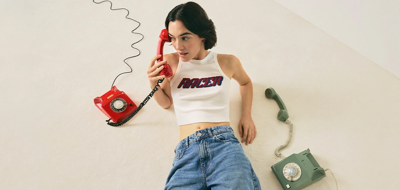 Donna in top corto bianco e jeans sdraiata sul pavimento, con in mano un telefono a rotella rosso, e accanto a lei un telefono a rotella verde.