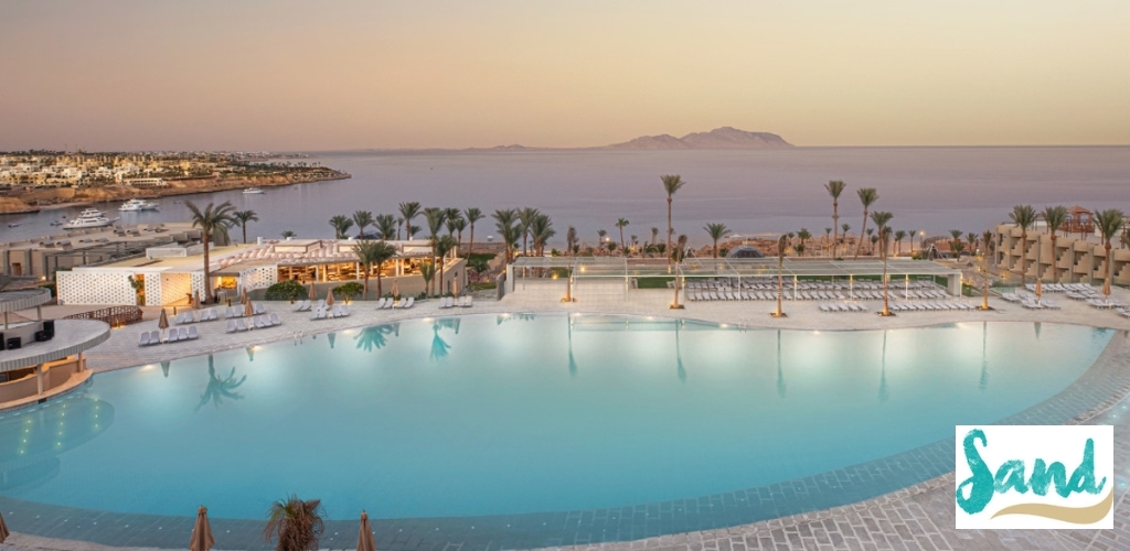 Sunset view of a large resort infinity pool with palm trees, sunloungers and yachts overlooking the calm sea and a distant island