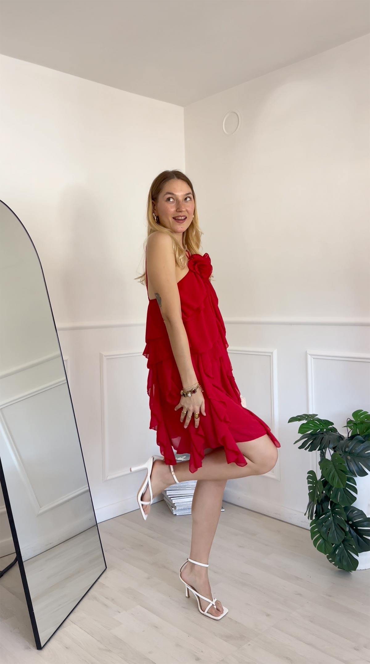 Woman in red ruffled dress and white heels posing by floor mirror and houseplant in minimal white room, smiling