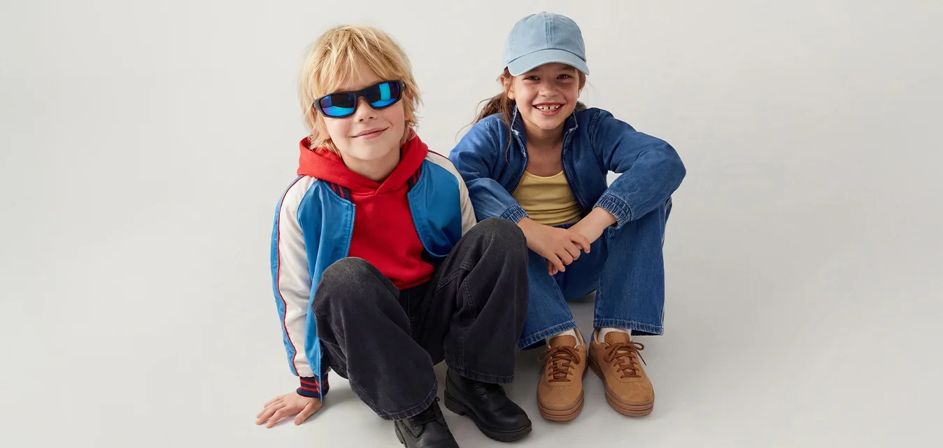Due bambini seduti per terra, uno con occhiali da sole e felpa rossa con cappuccio, l’altro con giacca blu e cappello, entrambi sorridenti.