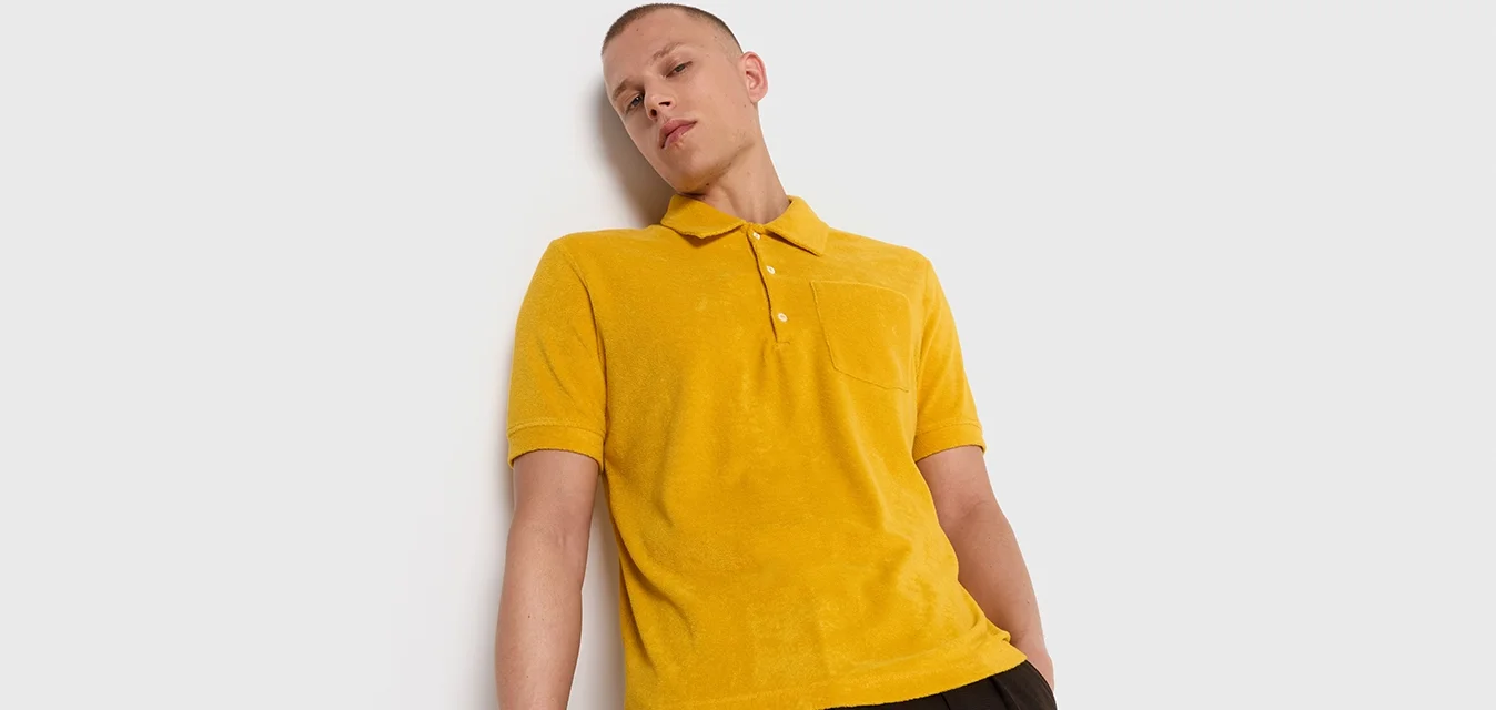 Polo giallo con una texture morbida, maniche corte e una pattina con tre bottoni. Presenta una tasca sul petto. Abbinato a pantaloni scuri larghi.