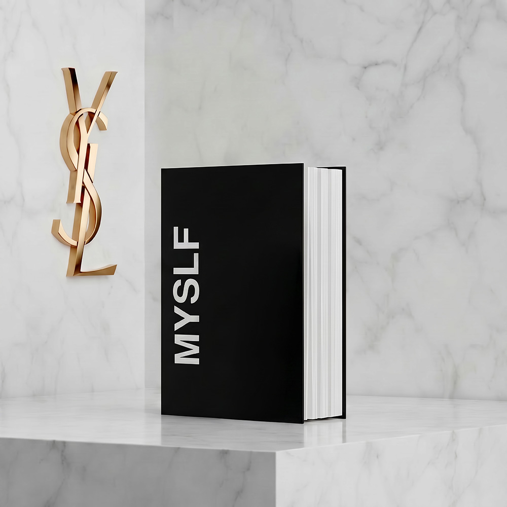 Un libro nero intitolato "MYSLF" esposto su un piedistallo di marmo bianco accanto a un logo YSL dorato.