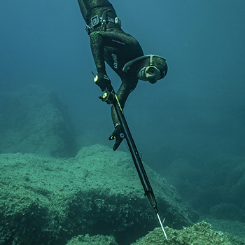 Freediver in wetsuit aiming a speargun at rocky seabed during underwater spearfishing in clear blue sea