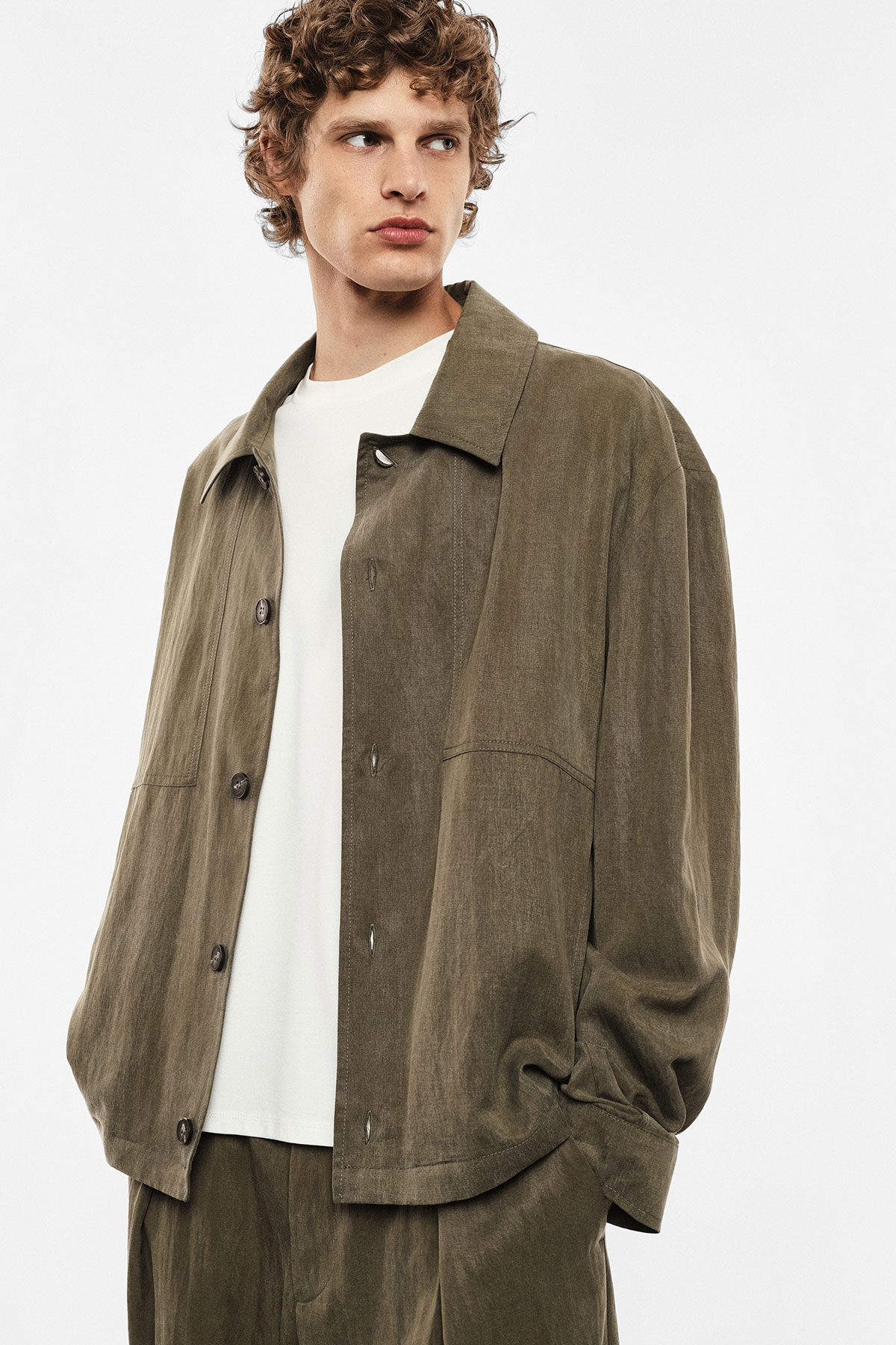 Young man wearing an olive oversized jacket and white T-shirt, hands in pockets, looking to the side on a white background