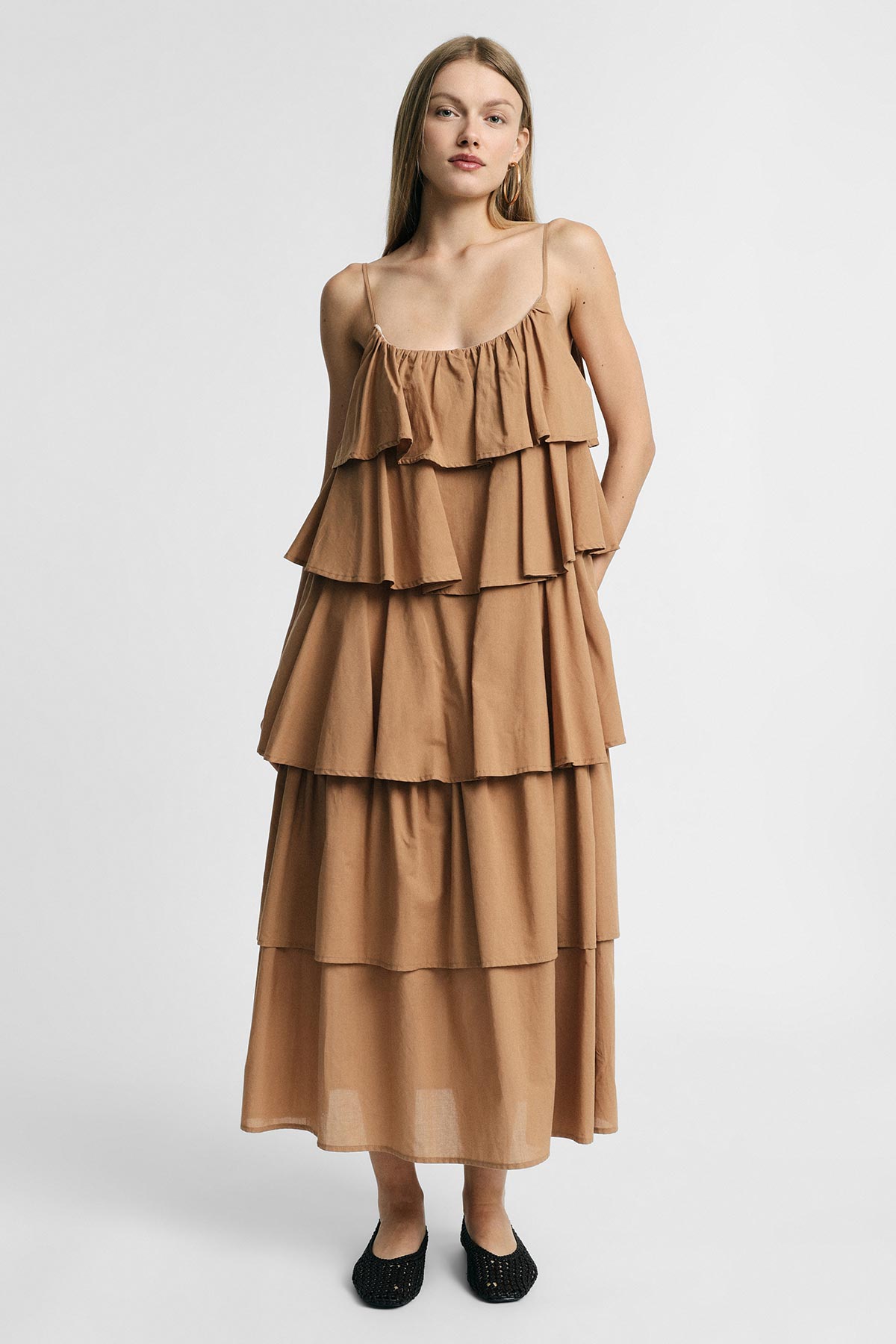 Model wearing camel tiered maxi dress with spaghetti straps and pockets, standing in black flats against light background