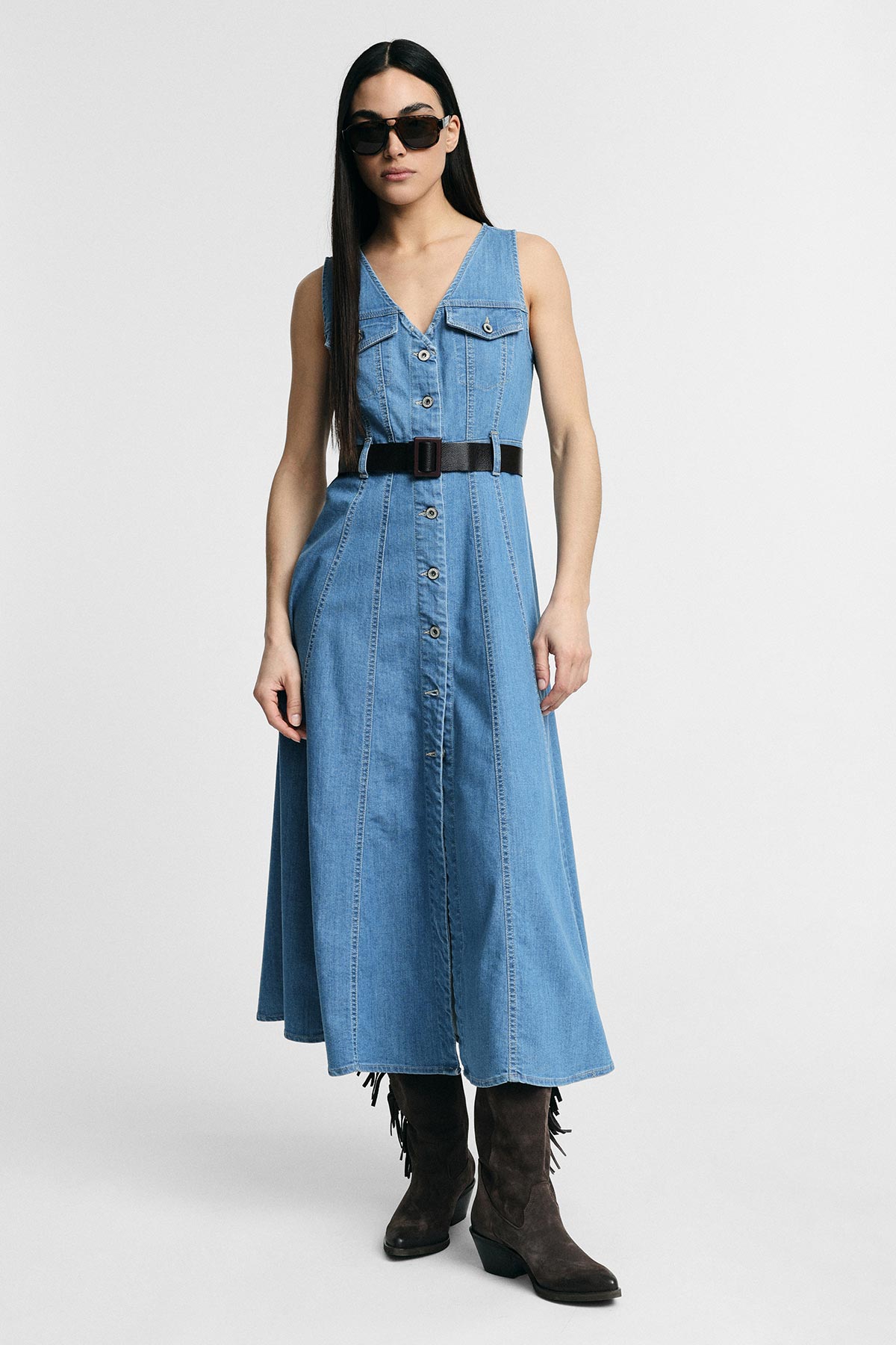 Model wearing sleeveless button-front denim midi dress with black belt, sunglasses and brown suede boots on a plain studio background