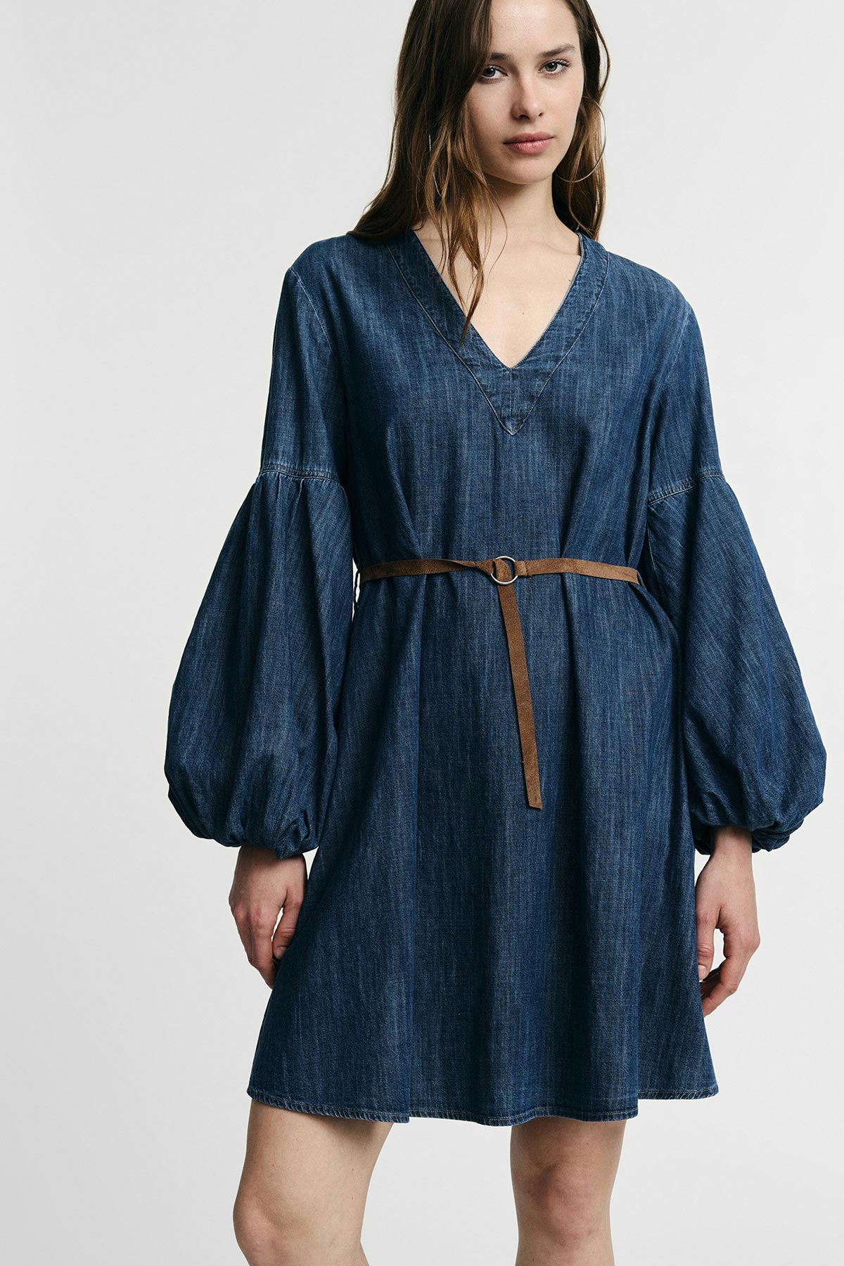 Woman wearing blue denim V-neck dress with puffed balloon sleeves and slim brown belt against a plain light background