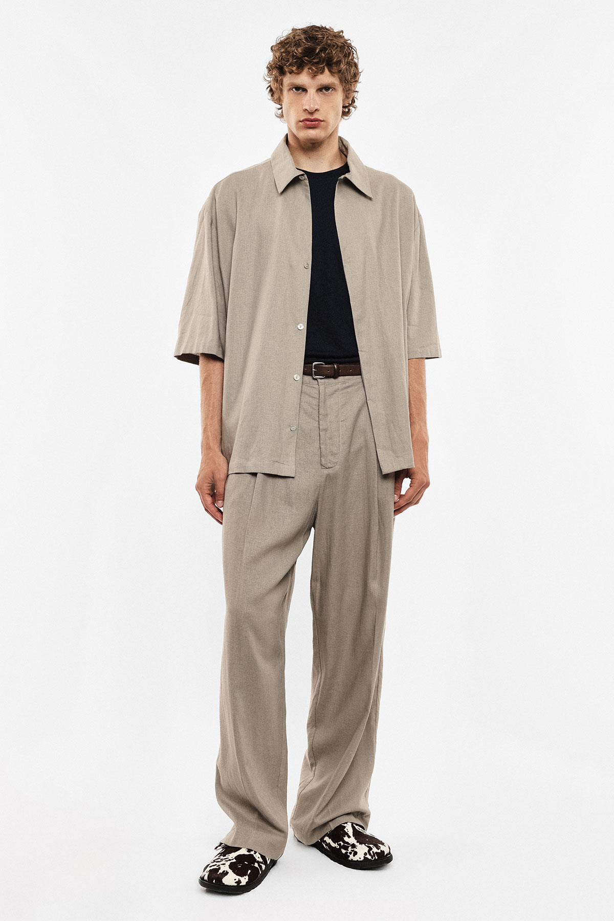 Male model wearing beige short-sleeve overshirt, black t-shirt, loose beige trousers and patterned slip-on shoes on white background