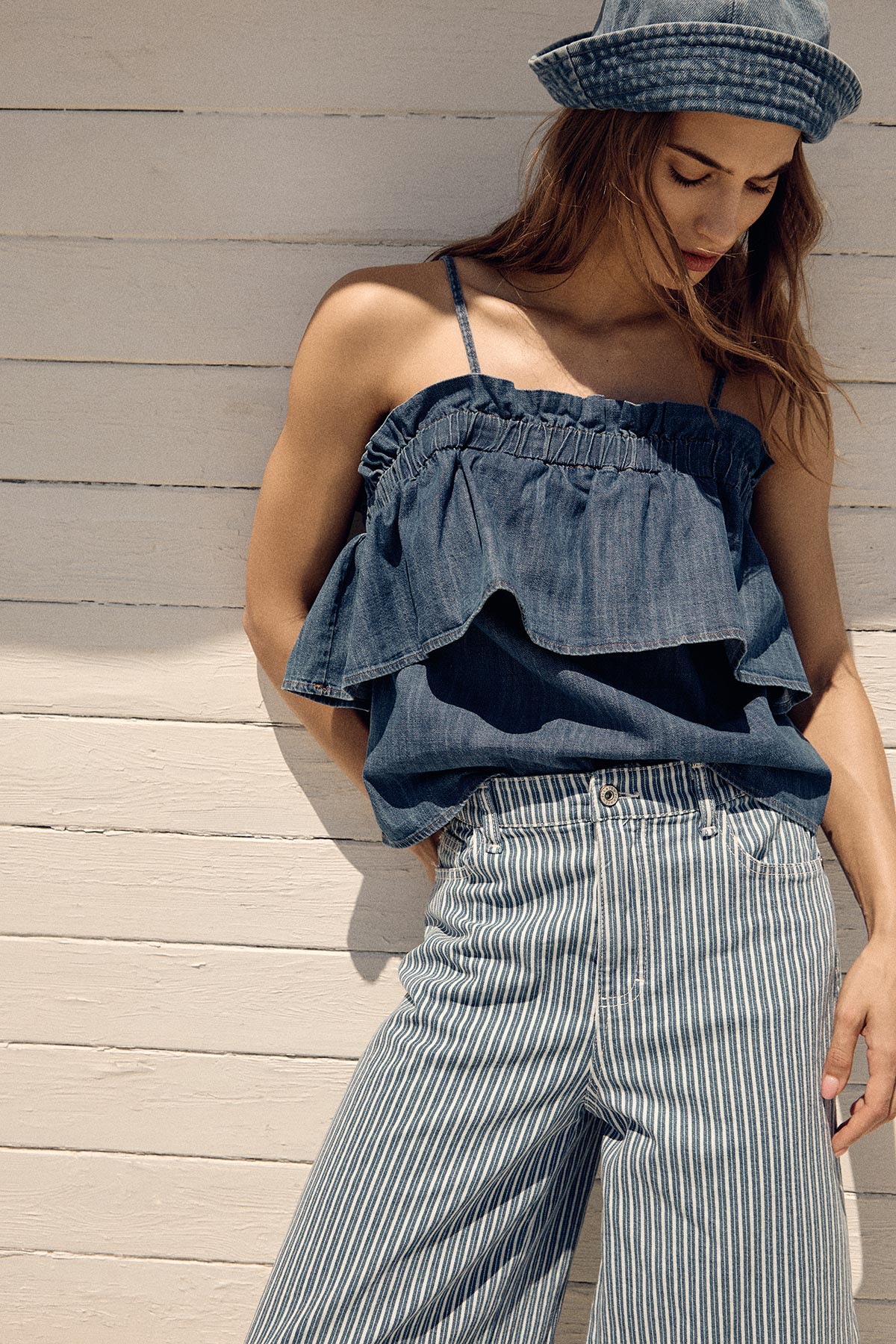 Woman in a denim ruffle cami top, striped high-waist trousers and denim hat leaning against a white painted wooden wall