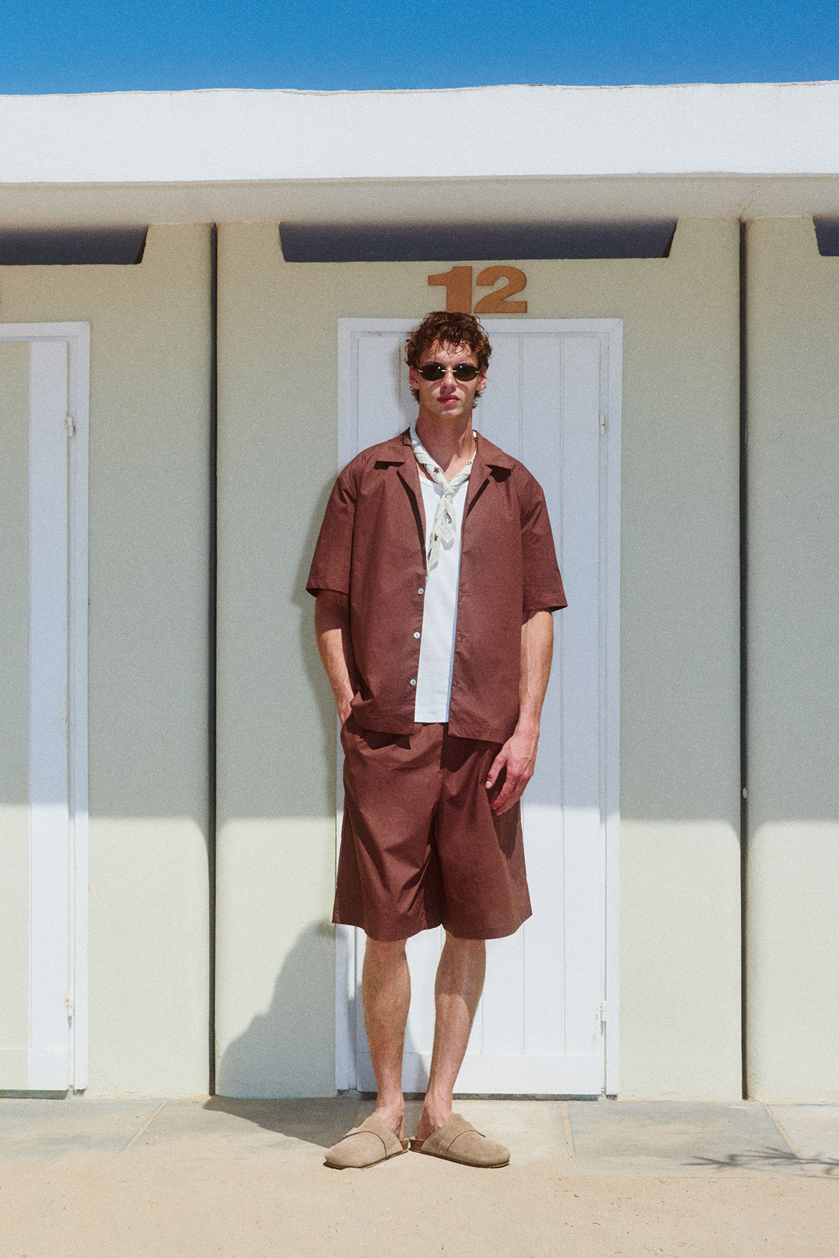 Man in brown matching summer co-ord and sunglasses standing in front of a white beach hut door numbered 12 in bright sunlight