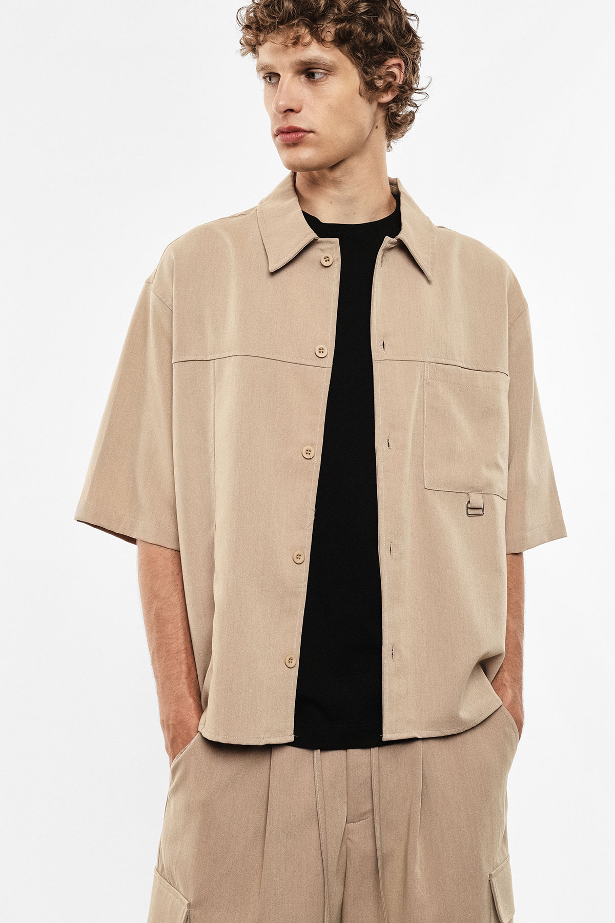 Man wearing beige short-sleeve button-up shirt and matching trousers over black T-shirt, hands in pockets, studio fashion shot