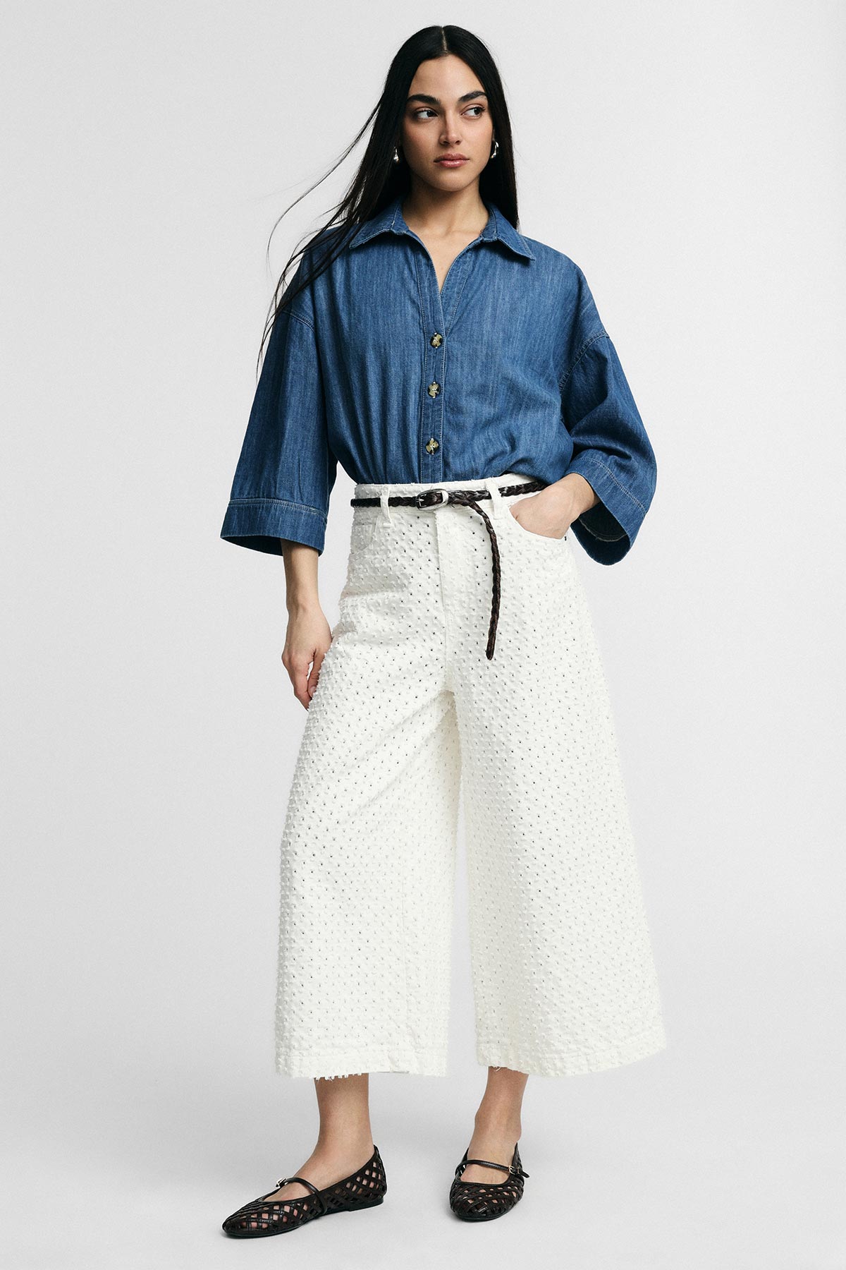Model wearing denim button-up shirt with white eyelet wide-leg trousers, braided belt and black woven flats, standing