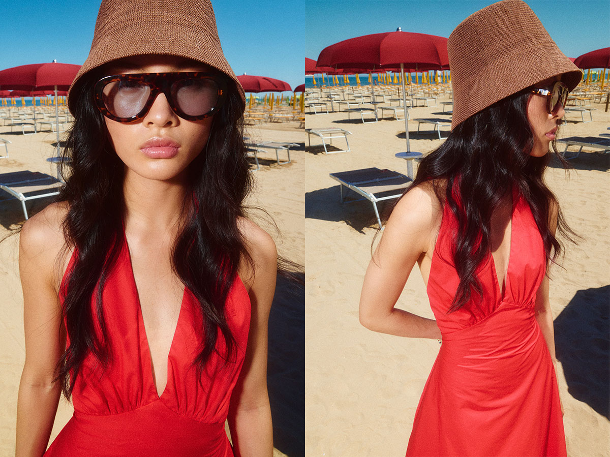 Woman in red halter dress, brown bucket hat and oversized sunglasses on sandy beach with red umbrellas and sunbeds