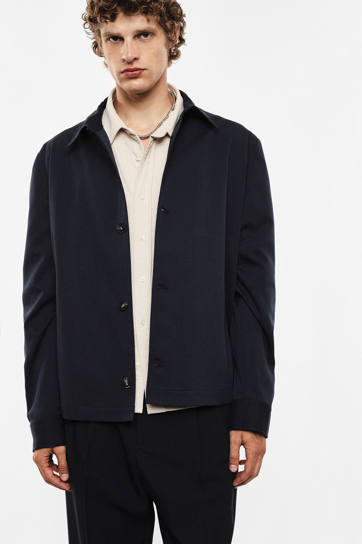 Male model wearing a navy button-up jacket over a beige shirt and black trousers, standing against a plain white background