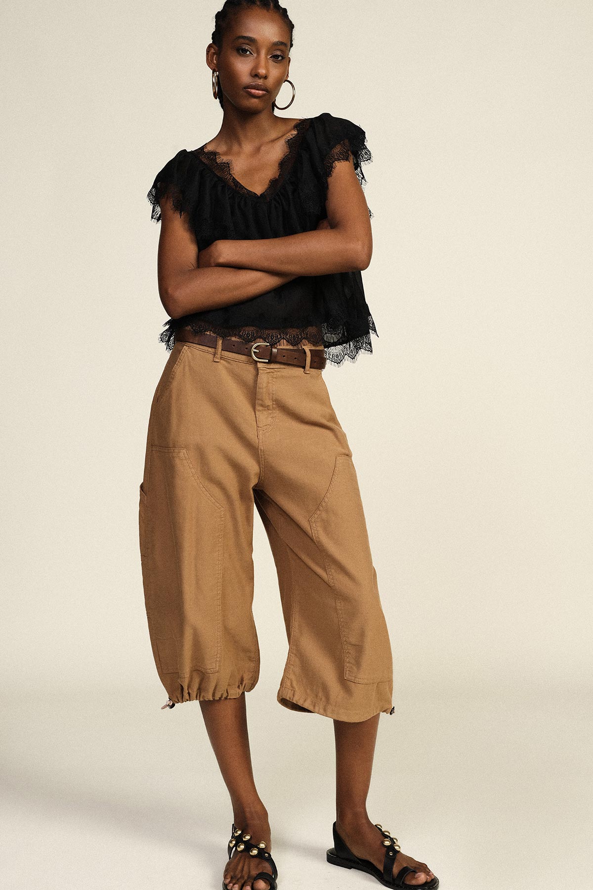 Woman wearing black lace-trim blouse, tan cropped wide-leg trousers, belt and embellished sandals, standing with arms crossed