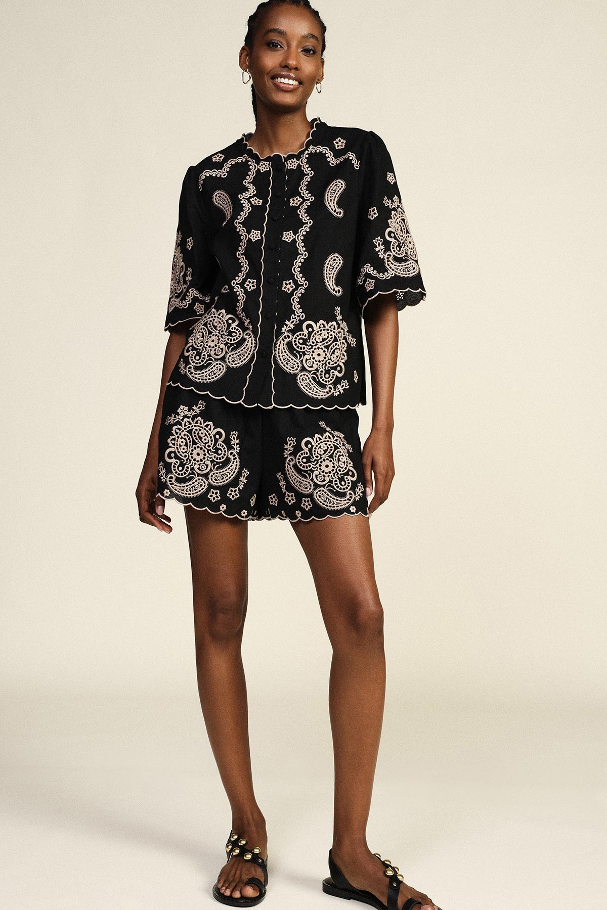 Smiling woman standing in a black embroidered two-piece set with scalloped edges and paisley motifs, wearing black sandals