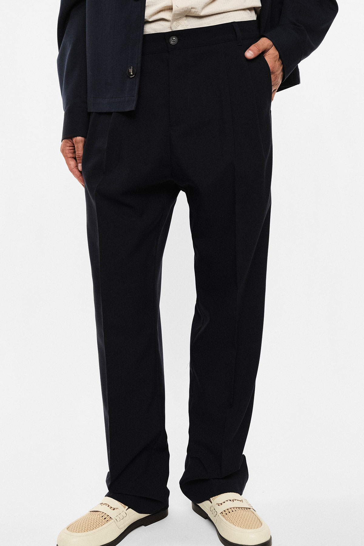 Tailored black men's trousers with front crease, hands in pockets, paired with cream loafers and navy jacket hem visible