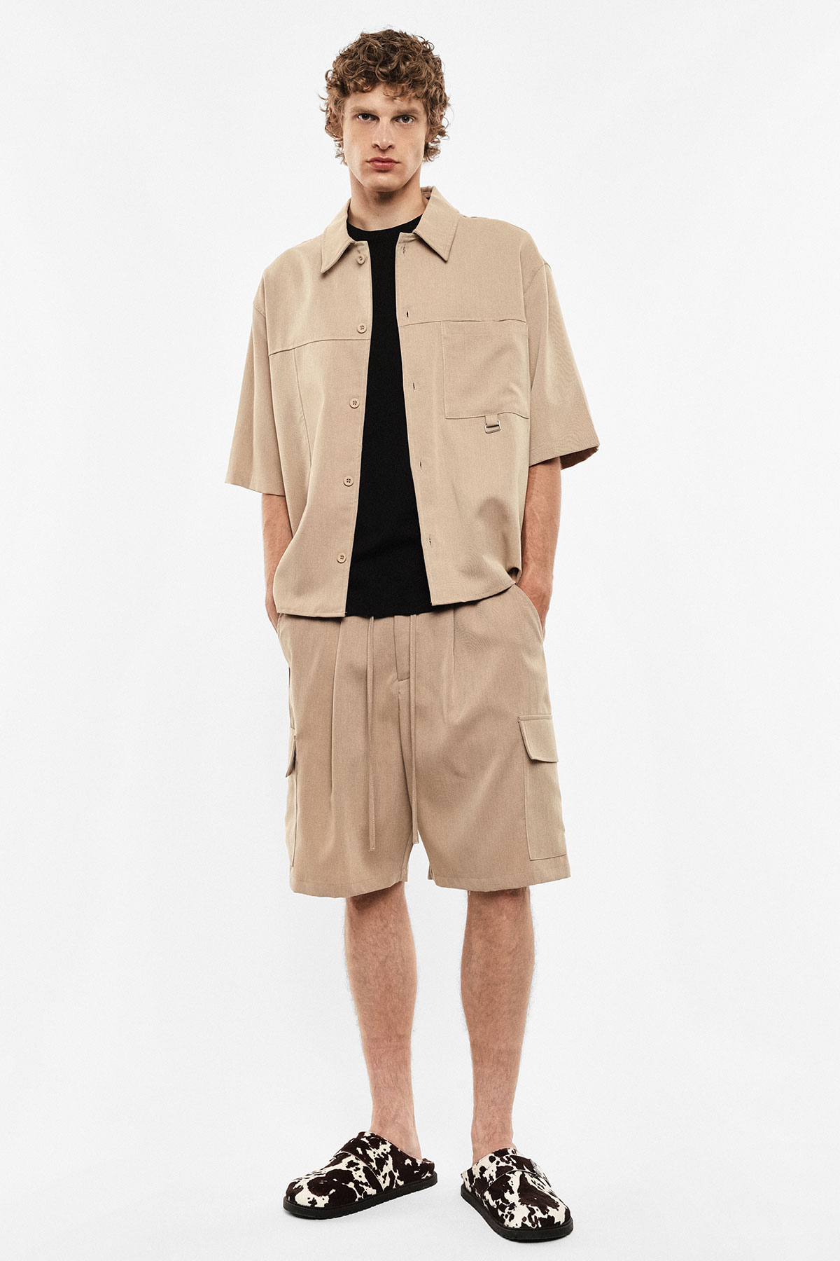 Male model wearing beige short-sleeve overshirt and matching cargo shorts with a black T‑shirt and patterned slip-on shoes