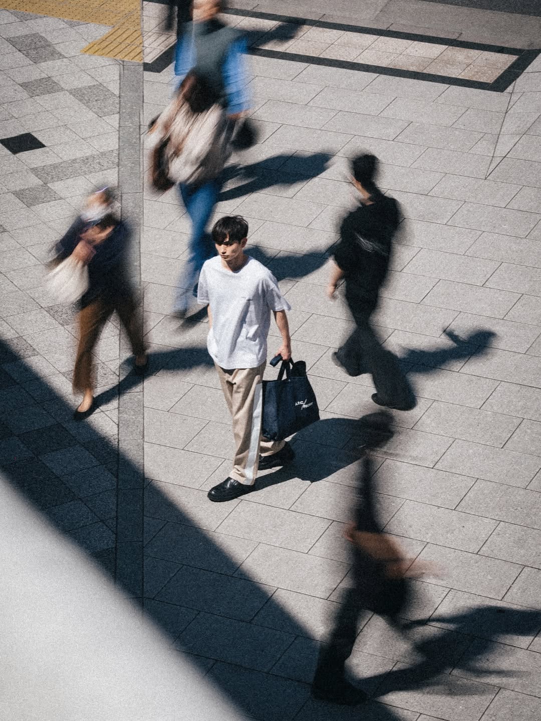 Young man in white t-shirt carrying a navy tote bag walks across a sunlit urban plaza amid blurred pedestrians and long shadows