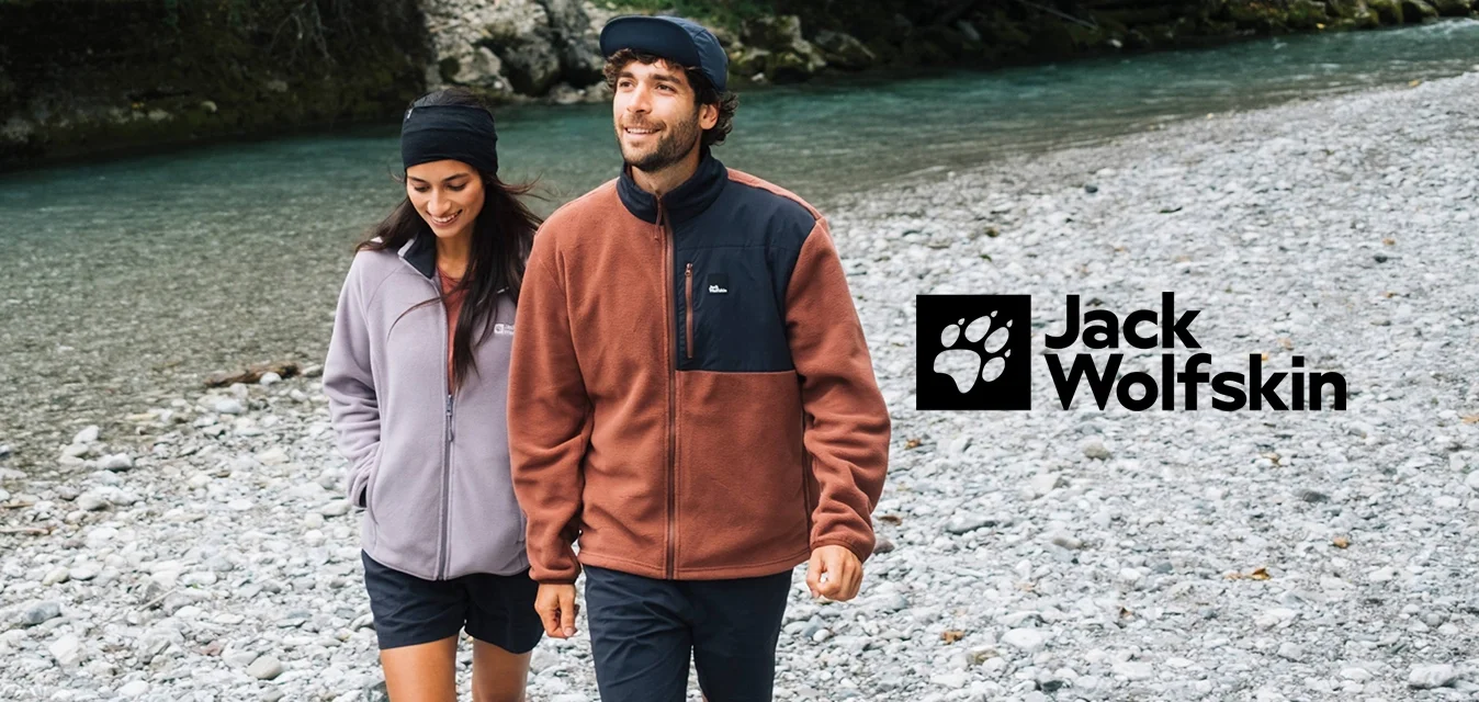 Coppia che passeggia su una riva rocciosa. Donna con giacca in pile lilla chiaro, uomo in pile marrone con pannello nero. Entrambi in pantaloni corti e sneaker.